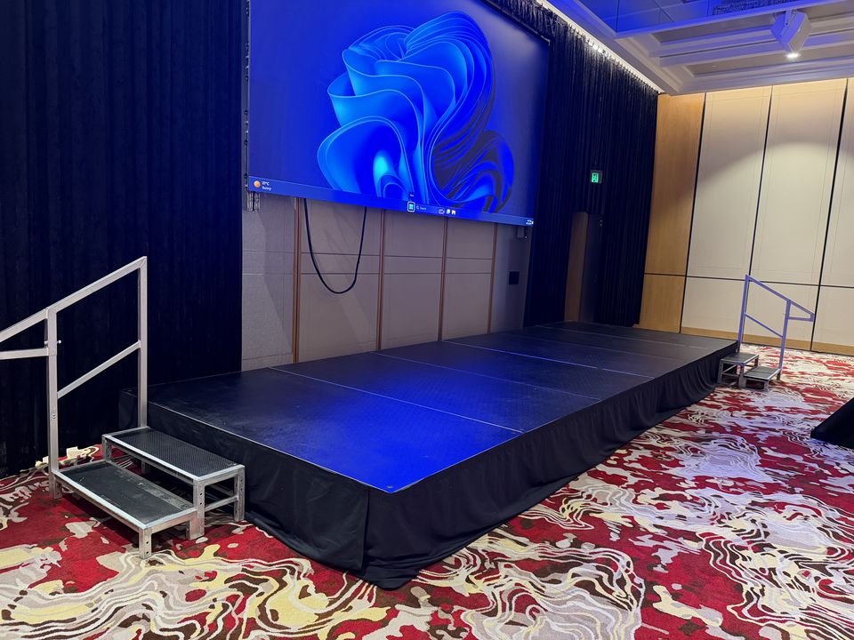 Black-skirted stage with handrailed steps in front of an LED screen in a function room