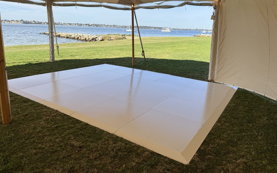 White dance floor on grass inside a sailcloth tent, with the river and moored boats behind