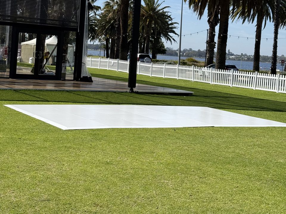 White dance floor on a riverside lawn, with palm trees and a white picket fence behind