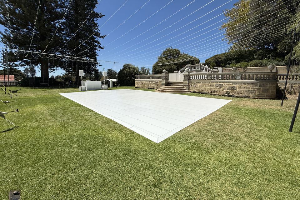 White dance floor laid on a garden lawn under festoon wires, with a stone balustrade and pine trees behind