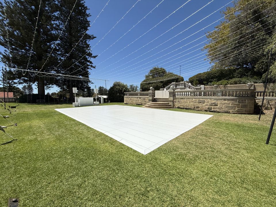 White dance floor laid on a garden lawn under festoon wires, with a stone balustrade and pine trees behind