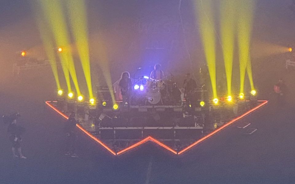 Triangle stage from CorpraHire's Wolfmother stage project, outlined in orange LED lighting, with a band performing under yellow spotlight beams in a darkened stadium