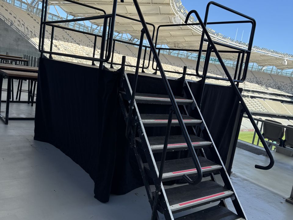 Aluminium steps with handrails leading up to a black-skirted, railed platform in a stadium stand
