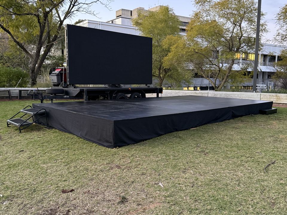 Black-skirted outdoor stage with a step unit on grass, with a mobile LED screen behind
