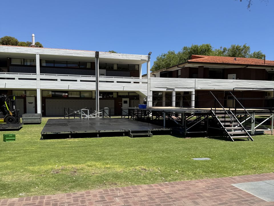 Two outdoor stages on a school lawn, one raised on scaffold legs with a stair unit
