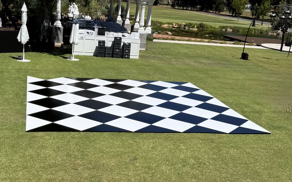 Black and white diamond-pattern dance floor laid on a lawn at a garden venue, with white columns, trees and a pond behind